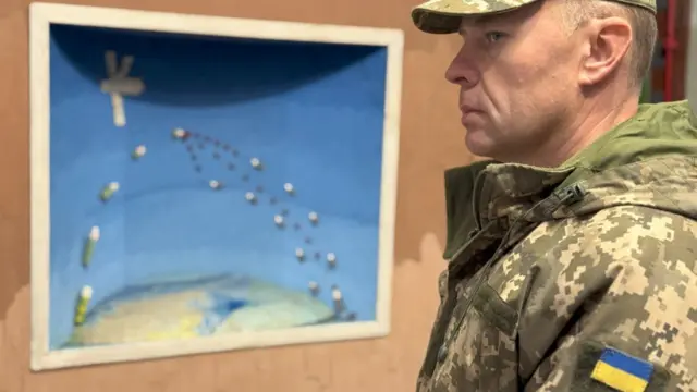Oleksandr Sushchenko vestido de militar ante un gráfico que muestra cohetes lanzados desde la Tierra