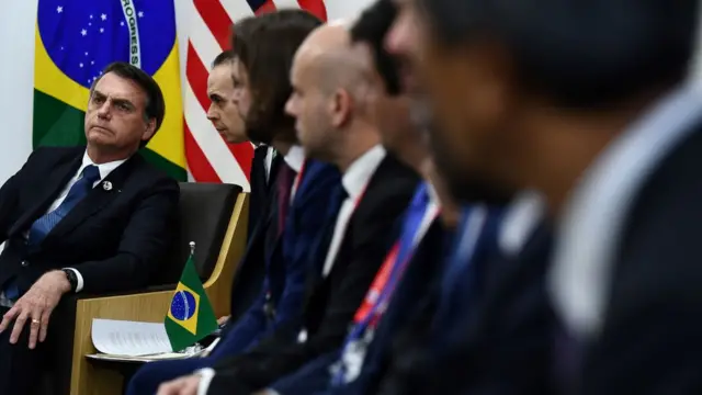 Bolsonaro aparece sentado em meio a outros homens engravatados e bandeiras