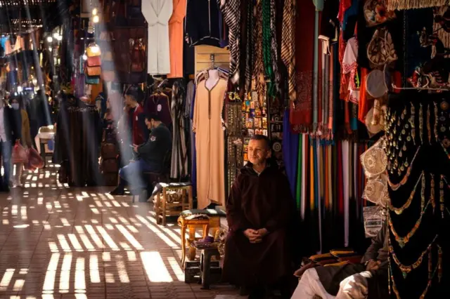 A man sits next to an market stall, with shafts of light piercing through the roof.
