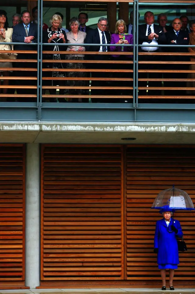 La reina Isabel II se resguarda de la lluvia mientras los invitados se refugian en la inauguración de la nueva sede de Lawn Tennis Associations en Roehampton, Londres, 2007