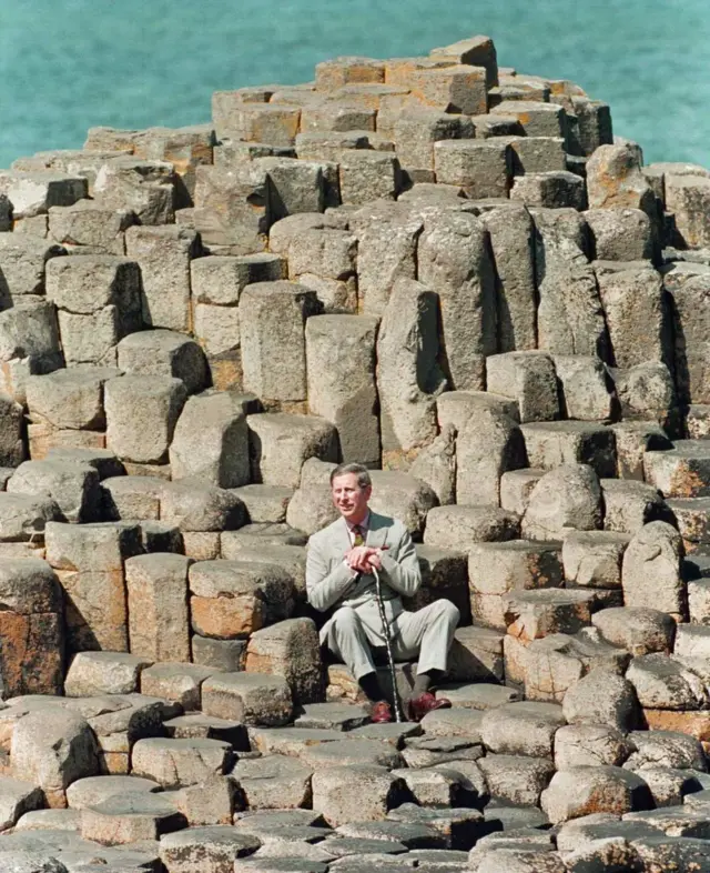 O príncipe Charles aparece sentado sozinho sobre pedras que compõem a Calçada dos Gigantes, um monumento natural na Irlanda do Norte