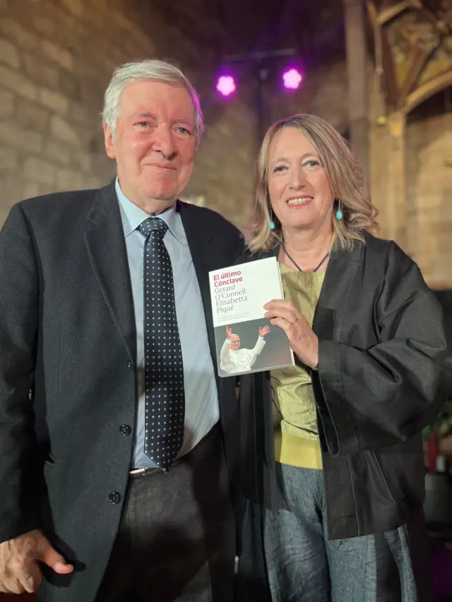 Jornalistas Elisabetta Piqué e Gerard O'Connell usam roupas social e seguram livro que estão lançando