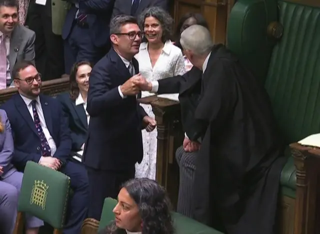 英下院で大勢が座る中、スーツ姿のバーナム氏が議長席の前に立ち、黒い議長服を着た議長と握手している
