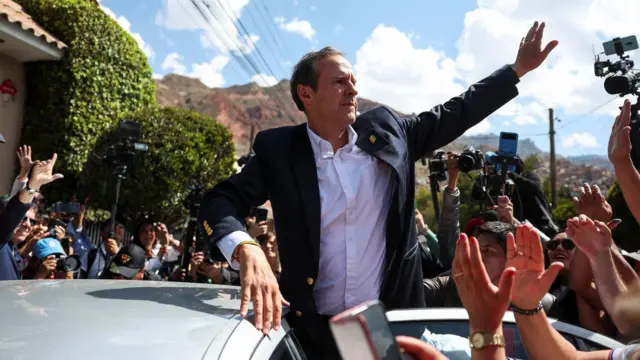 Jorge Tuto Quiroga, con chaqueta oscura y camisa blanca, saluda desde lo alto de un auto. Se ven manos su alrededor alrededor, cámaras de fotos y, de fondo, una montaña