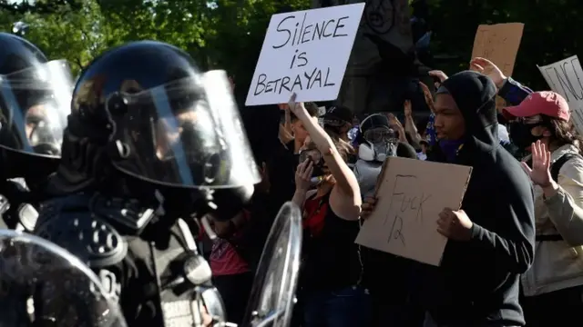 Manifestantes com cartazes protestam em frente a grupo de policiais com armas e equipamentos anti-manifestação