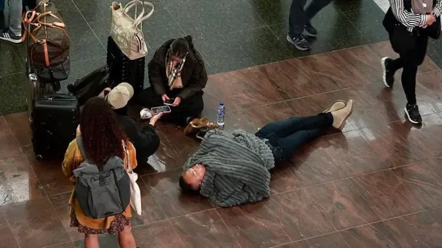 毛布にくるまって床で寝る男性や、座り込んでスマートフォンを操作する女性