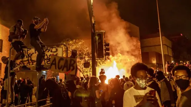 Protesto contra racismo nos Estados Unidos, com uma área pegando fogo e pessoas com cartazes