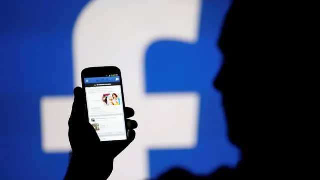 A man is silhouetted against a video screen with an Facebook logo as he poses with an Samsung S4 smartphone in this photo illustration August 14, 2013