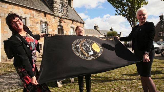 Ativistasroleta virtual de numerosCulross, Escócia, segurando uma bandeira que diz: "Lembrando as bruxas acusadas da Escócia"
