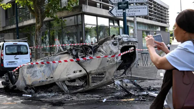 A pedestrian photographs an overturned burnt car in Aubervilliers, north of Paris following riots on 30 June 2023