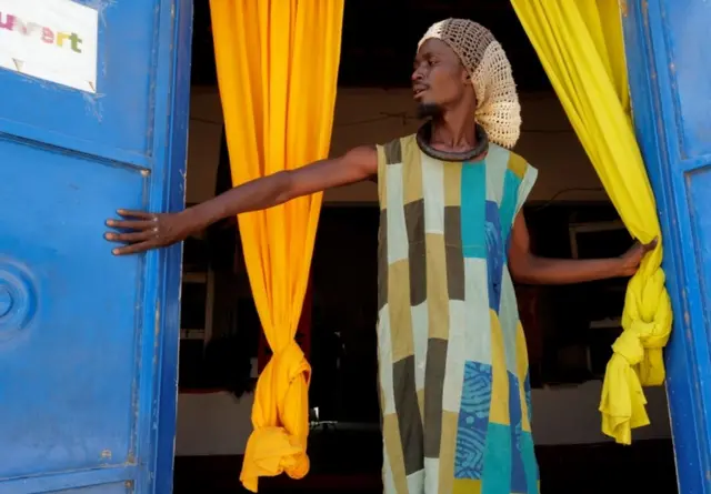 A man stands at the entrance to a community shop. He is wearing bright clothes that match the curtains and doors.