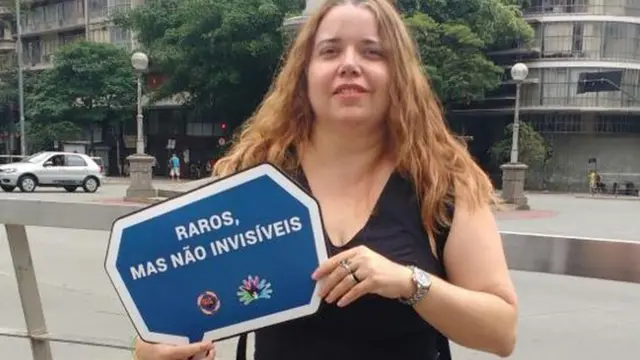 Margareth na rua, sorrindo e com placa na mão que diz: 'Raros, mas não invisíveis'