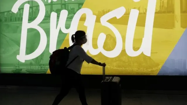 Mulher caminha com mala em frente a propaganda do Brasil
