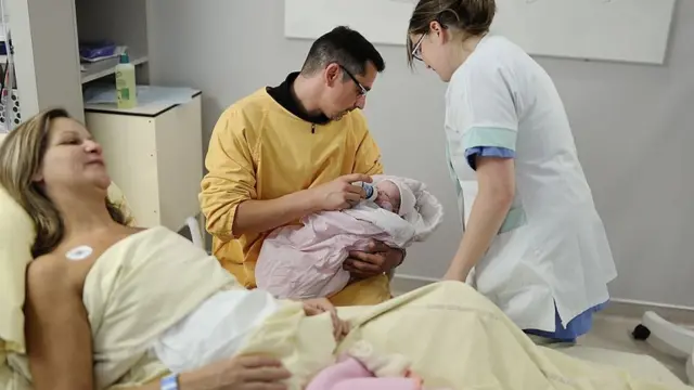 Un hombre da un biberón a un bebé recién nacido junto a la cama donde se encuentra la madre.