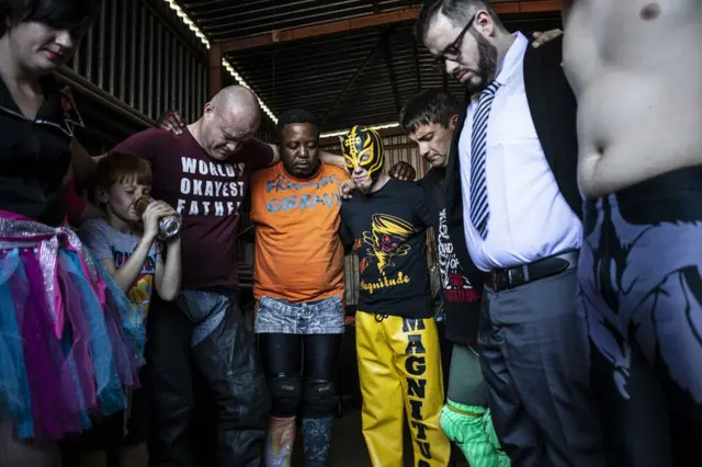 Members of a wrestling community are seen in prayer with their arms around each other.