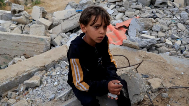 طفل في خان يونس يرتدي بدلة رياضية سوداء وأكمامها مخططة باللون الأصفر والأبيض، يجلس على ردم مبنى مهدم