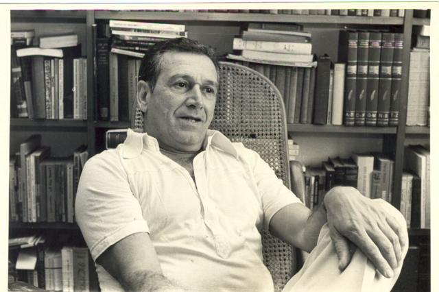 Sabino sentado e falandoaposta caixa appfrente a estantesaposta caixa applivros