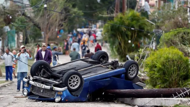 Un auto dado vuelta en el medio de la calle en La Habana mientras personas toman fotografías.