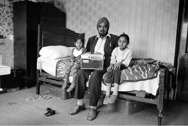 
Picha nyeusi na nyeupe ya mwanamume akiwa amekaa mleleoni wa kitanda akiwa na binti zake wawili . Ameshikilia redio aliyoipakata mapajani.