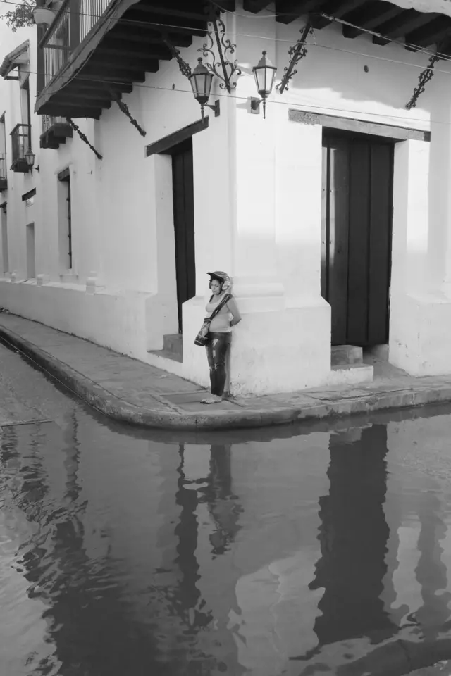 Mujer esperando en una esquina inundada tras una lluvia y su reflejo en el agua.