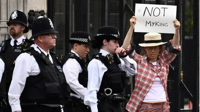 Fotografia colorida mostra uma pessoa branca de óculos segurando um cartaz escrito "not my king", ou "não é meu rei" em inglês, ao lado de pessoas com fardas (em frente ao palácio de Westminster).