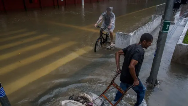 Jovem pedala em meio a alagamento em São Paulo, enquanto homem tenta transportar carroça