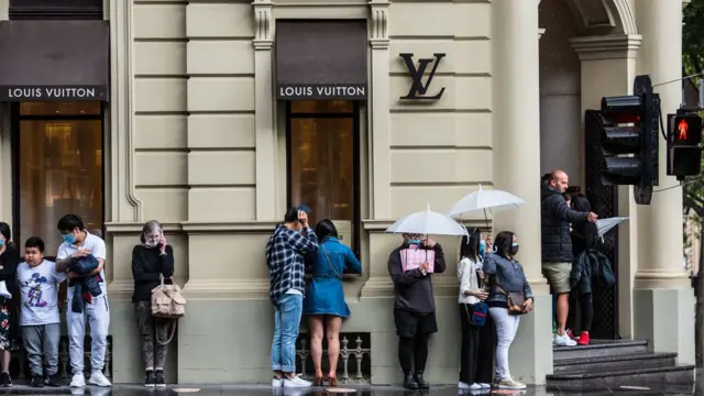 लोग बारिश में खड़े होकर लुइ विटन स्टोर में घुसने का इंतज़ार करते हुए