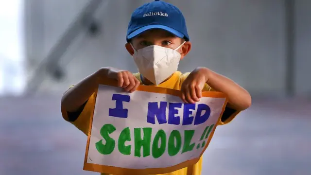 Un niño sostiene un cartel con la frase en inglés: "I need school" (necesito escuela)