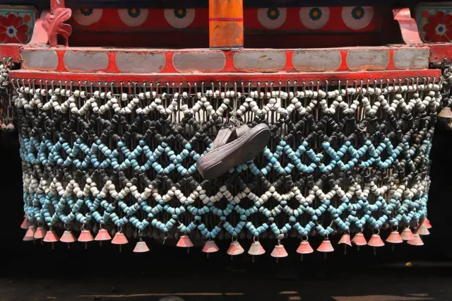 Detalj prednjeg branika Bedford kamiona u Pešavaru, Pakistan sa kojeg visi niz lanaca sa zvončićima, kao i dečija cipela. Na vozila se postavljaju različiti predmeti i simboli koje imaju funkciju amajilja i talismana koji štite od zlih sila: među njima su životinjski rogovi, repovi jaka, antene u obliku bumeranga, trake sa sufijskih grobnica, lanci, zvončići i cipele.