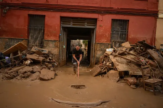 Un hombre saca barro de una casa inundada. Fuera hay un montón de muebles y enseres embarrados.