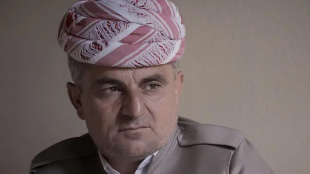 صورة لرجل في منتصف العمر يرتدي غطاء على رأسه وسترة بنية اللون وينظر بتأمل.