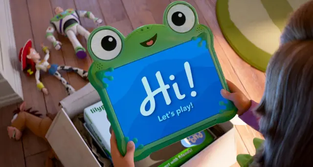 Niña sostiene una tablet verde con forma de rana que muestra “Hi! Let’s play!” ("¡Hola! ¡Juguemos!") mientras juguetes están esparcidos en el suelo.
