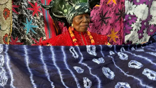Mama Nike holds a large piece of dark blue patterned adire cloth, wearing a bright red beaded garment, a large green patterned headwrap and a necklace of red and orange beads. She looks down at the cloth. Behind her is a display of vividly coloured textiles in black, pink, purple and white, featuring floral and star-like designs that fill the background.