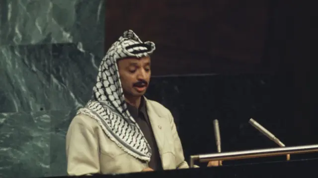 ياسر عرفات، مرتدياً الكوفية الفلسطينية وبزة عسكرية، يلقي خطاباً أمام الجمعية العامة للأمم المتحدة في نيويورك عام 1974