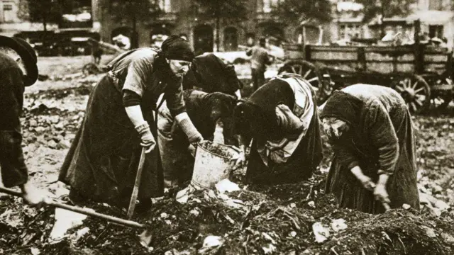 চারজন মহিলা ও একজন পুরুষ কোদাল ও বেলচা হাতে আবর্জনার স্তূপে ঝুঁকে কাজ করছেন। মহিলাদের মধ্যে একজন ব্যবহার্য জিনিসপত্র সংগ্রহের জন্য একটি বালতি ধরে আছেন।