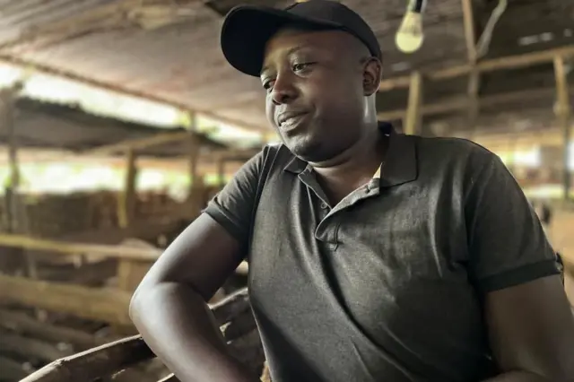 Olivier Gaston Hagenimana yambaye ingofero y'umukara n'umupira wijimye ujya kuba umukara. Ahagaze iruhande rw'ikiraro cy'ingurube, arareba hasi ku ruhande, hejuru ye ku gisenge cy'inzu hari itara ridacanye. 