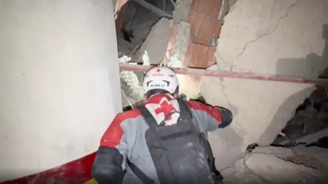 Рятувальник у захисному спорядженні, який пробирається крізь вузький прохід серед завалів зруйнованої будівлі. Навколо – потріскані бетонні конструкції, уламки цегляної кладки та пошкоджені стіни зі значними тріщинами. Сцену освітлює штучне світло, ймовірно ліхтар або прожектор.