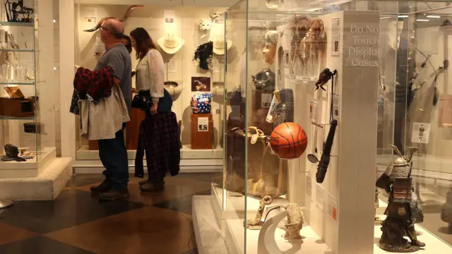 Відвідувачі у музеї роздивляються експонати за склом - видно різні речі, капелюхи, перуки, баскетбольний мʼяч тощо