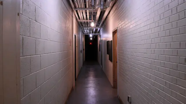 A long industrial hallway, with the walls made out of concrete blocks painted white and a cement floor. Pipes are visible running along the ceiling. 