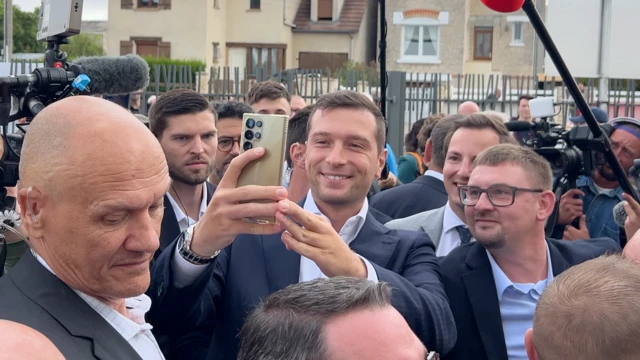 Jordan Bardella prend un selfie au milieu d'une foule de journalistes et de partisans lors d'un rassemblement.