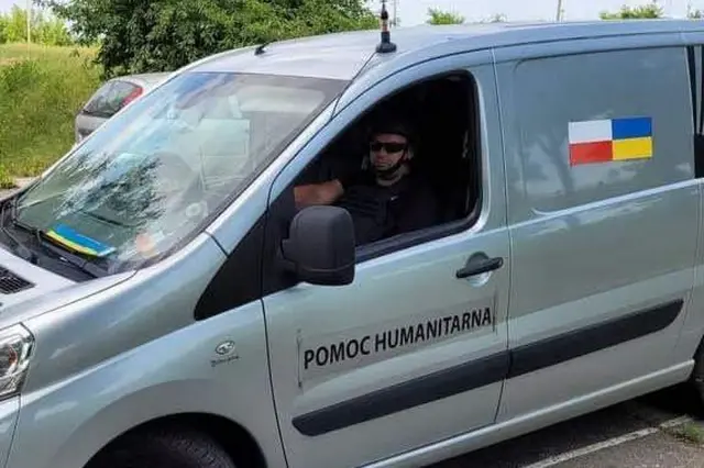 Сірий мікроавтобус із написом "POMOC HUMANITARNA" ("Гуманітарна допомога") та прапорами України й Польщі; за кермом сидить людина в темних окулярах.