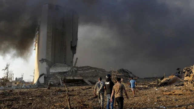 Três homens caminham em meio a fumaça e estilhaços, com prédio destruído atrás