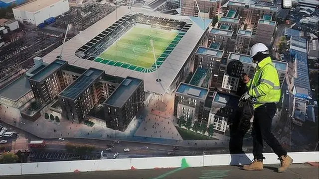 A construction worker walks past a hoarding displaying an impression of the completed new stadium at Plough Lane