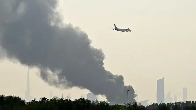 Avião da companhia aérea Emirates voa perto de colunas de fumaça de um incêndio perto do Aeroporto Internacional de Dubai, nos Emirados Árabes Unidos, no dia 16 de março de 2026