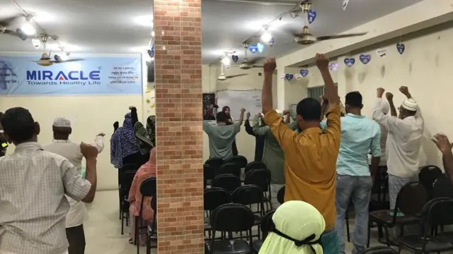 একজন নারী কর্মী মাইক ব্যবহার করে বেশ কয়েকজন বয়স্ক নারী-পুরুষকে ব্যায়াম করাচ্ছেন