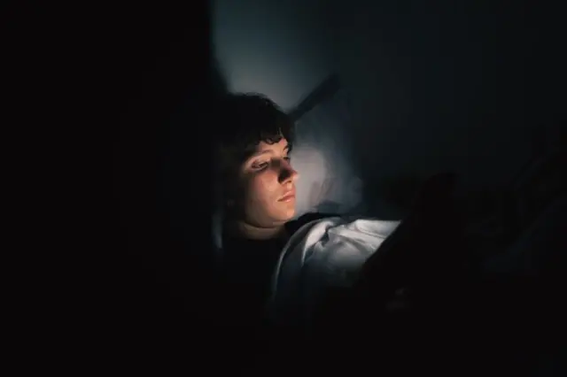 Gece yatağındaki gencin yüzüne telefon gibi bir ışık vuruyor. 
