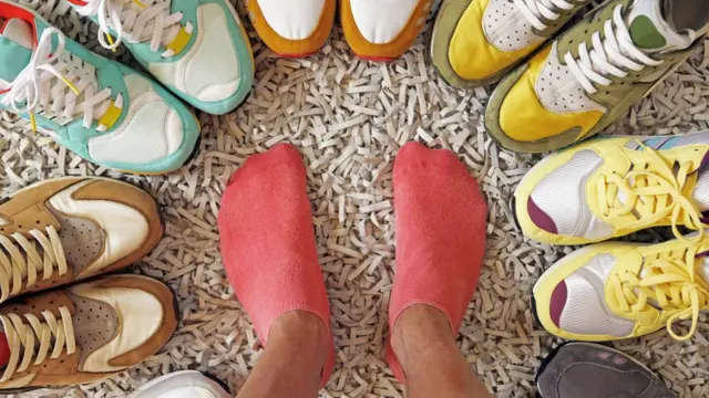 Unos pies rodeados de pares de zapatos distintos.