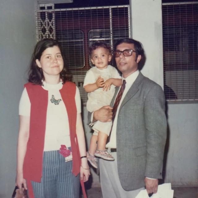 Foto de Mithu Sanyal quando criança; seu pai veste terno cinza e a carrega com uma das mãos; ao seu lado, sua mãe de colete vermelho e calça azul