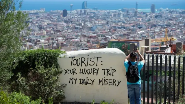 Um homem fotografa a paisagemroda roletaBarcelona, ao ladoroda roletauma parede onde está escrito: 'Sua viagemroda roletaluxo, minha desgraça diária'