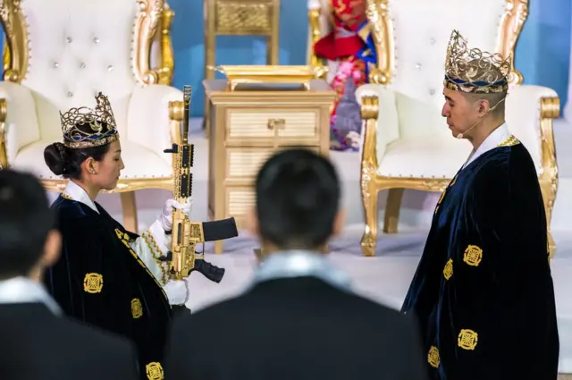 القس هيونغ جين مون يستعد لأخذ بندقية ذهبية من طراز AR-15 التي ترمز إلى عصا الحديد المذكورة في الإنجيل من زوجته الكاهنة ييوون أه لي مون اللذين ينحدران من كوريا الجنوبية خلال احتفال باستمرار الزواج بين الأزواج بمناسبة المعبد المقدس للسلام والتوحيد العالمي وهو جزء من كنائس التوحيد في نيوفواندلاند في بنسلفانيا بالولايات المتحدة.